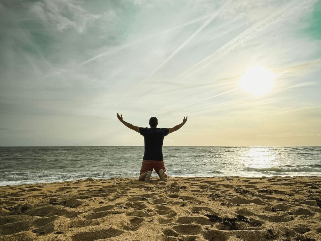 homme, à genoux sur la plage, face à la mer et les bras levés en direction du sunset, pour exprimer sa gratitude 