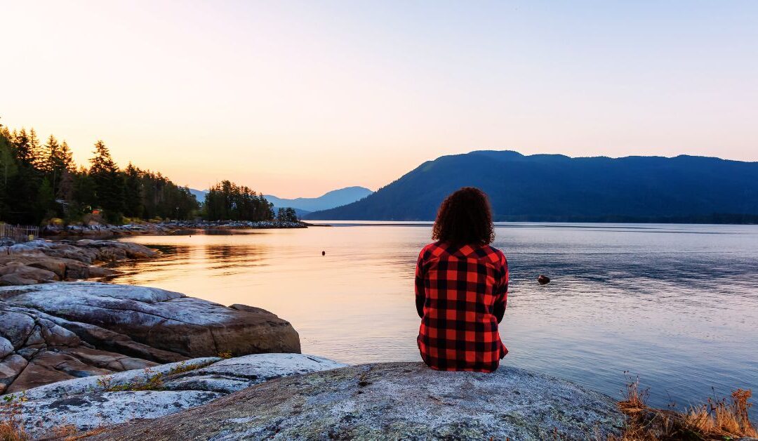 Femme assise sur un rocher face à un lac, au lever du soleil, pour se reconnecter à soi même
